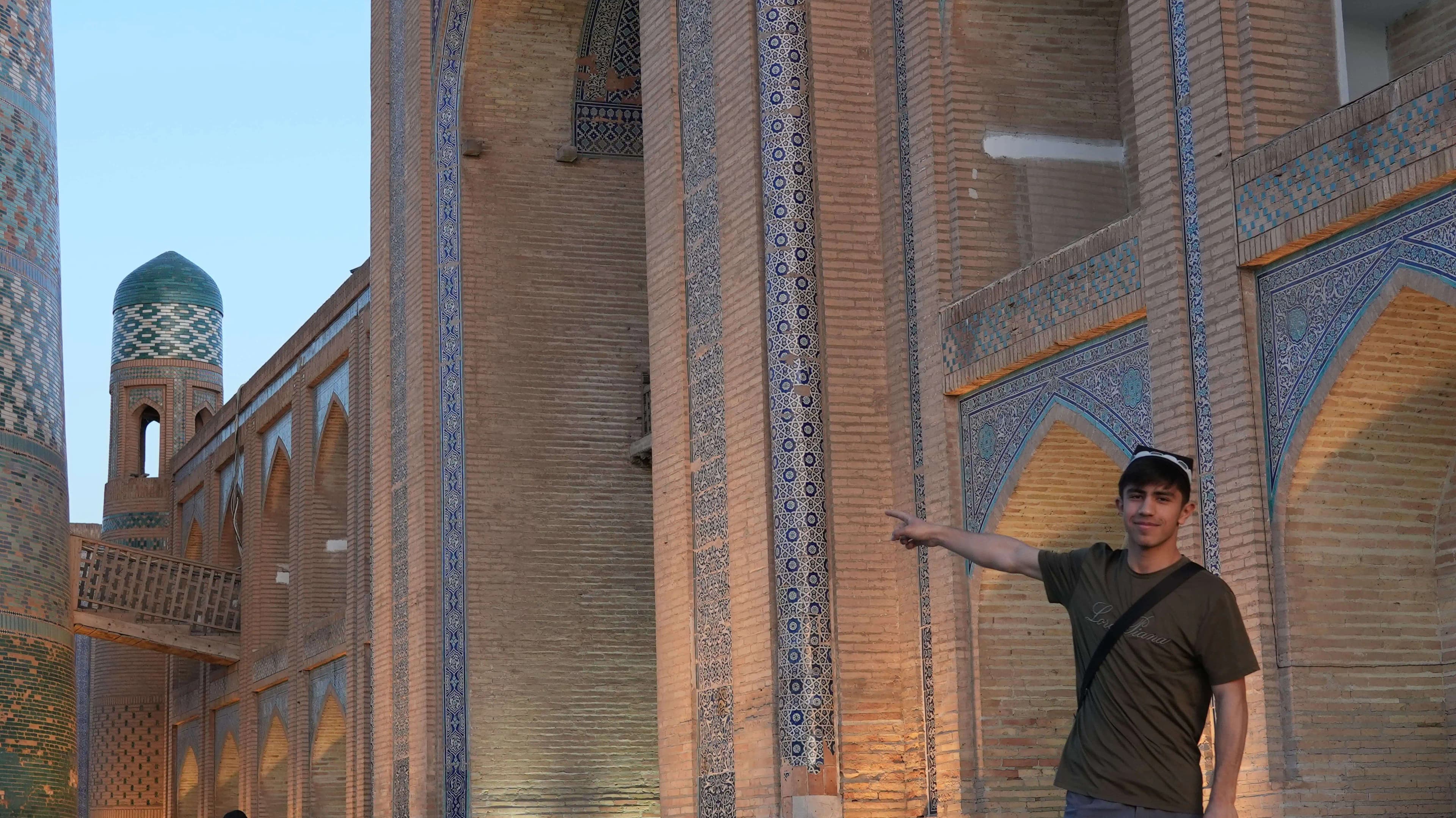A man in Khiva’s old town points out the blue tilework on a madrasa wall, with a tiled minaret behind him.
